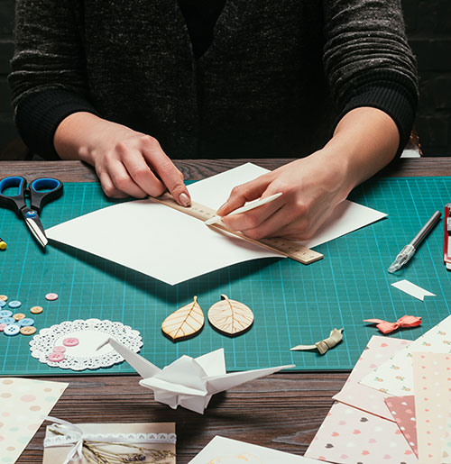 hands scoring fold of handmade card with decorative materials on work surface