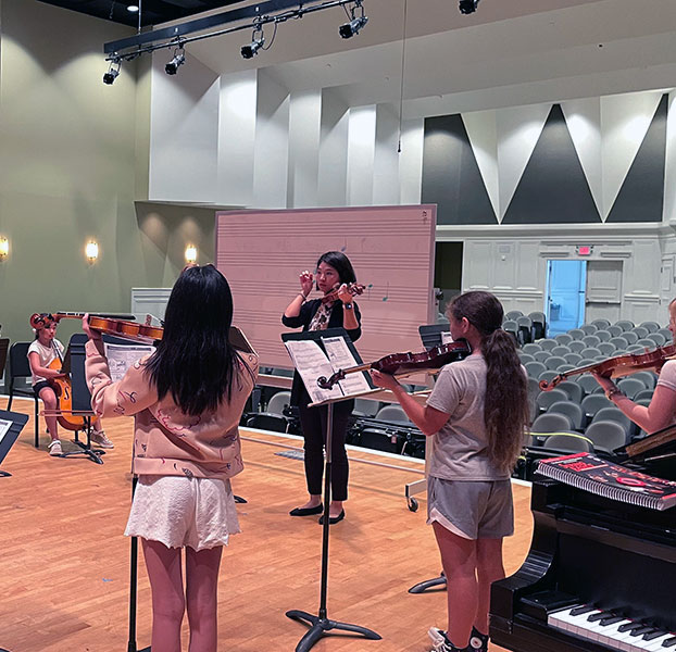 students in auditorium playing various string instruments with instructor